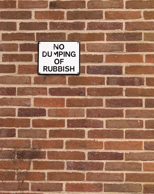 A rectangular metal sign mounted on a red brick wall features the message 'NO DUMPING OF RUBBISH' in bold black uppercase letters on a white background. The sign has a black border and is positioned slightly above the centre of the image, with visible mounting brackets at each corner. The brick wall is made of uniform, reddish-brown bricks with darker mortar joints, creating a textured surface that extends beyond the sign across the entire frame. The lighting is even, highlighting the rough texture of the brickwork and the smooth surface of the sign. This scene depicts a common external surface where waste management regulations apply, and the sign serves as a clear reminder against illegal dumping, aligning with services offered by Waste Disposal Lewisham in managing and preventing rubbish accumulation in residential or commercial areas.