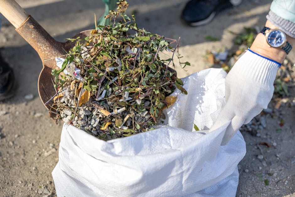 A person wearing a white glove and a wristwatch is holding a large white woven polypropylene bag filled with garden debris, including small branches, twigs, green ivy leaves, and dried foliage. The debris appears to be a mix of natural plant waste and some small, broken pieces of plastic litter. The bag is open at the top, with the plant material visibly overflowing from its rim. An orange-brown metal shovel with a worn, rusted surface is positioned on the left side of the image, angled as if in use for collecting or loading waste. The background shows uneven, bare ground with some loose gravel and dirt, indicating an outdoor setting suitable for garden clearance or rubbish removal activities. The scene suggests an organised effort for garden waste collection, possibly part of a private clearance or waste disposal service by Waste Disposal Lewisham, aligning with the context of rubbish removal and on-site clearance services.