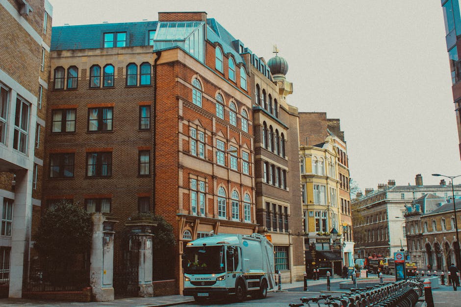A city street scene featuring a mix of historic and modern architecture, with multi-storey buildings constructed from red brick, beige stone, and painted facades. The buildings have large windows, some arched and others rectangular, with decorative features such as cornices and ornate trims. A prominent building in the center has an intricate design with multi-paned windows, a rounded turret with a greenish dome, and a small weather vane on top. The street is lined with parked vehicles, including a light blue waste collection truck labeled with the company name Waste Disposal Lewisham, positioned near the curb in the foreground. The pavement is made of concrete, and there are metal bike racks on the sidewalk, along with bollards separating the pedestrian area from the road. Street lamps and signage are visible further down the street, with overcast sky providing diffused lighting. The scene evokes a typical urban environment where independent waste collection services handle rubbish removal, evident from the presence of the waste disposal vehicle and the lack of visible council bins, supporting private and on-site clearance activities in a central London context.