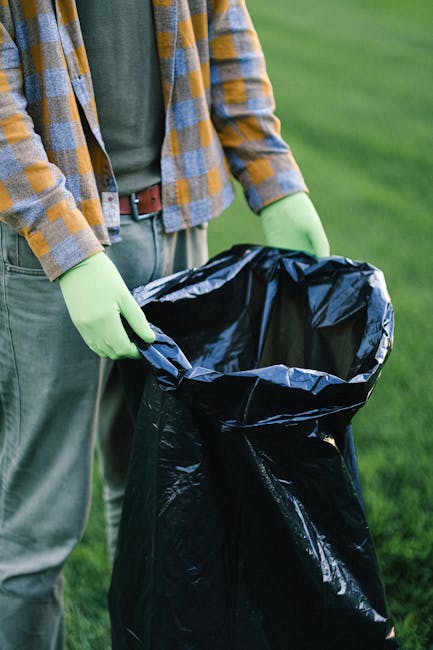 A person wearing a white glove and a wristwatch is holding a large white woven polypropylene bag filled with garden debris, including small branches, twigs, green ivy leaves, and dried foliage. The debris appears to be a mix of natural plant waste and some small, broken pieces of plastic litter. The bag is open at the top, with the plant material visibly overflowing from its rim. An orange-brown metal shovel with a worn, rusted surface is positioned on the left side of the image, angled as if in use for collecting or loading waste. The background shows uneven, bare ground with some loose gravel and dirt, indicating an outdoor setting suitable for garden clearance or rubbish removal activities. The scene suggests an organised effort for garden waste collection, possibly part of a private clearance or waste disposal service by Waste Disposal Lewisham, aligning with the context of rubbish removal and on-site clearance services.