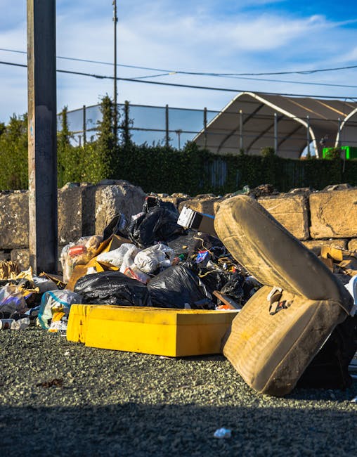 A close-up view of an outdoor area showing a pile of mixed rubbish including black and white plastic garbage bags, a yellow plastic container, and an old, dirty, and worn car tire leaning against a stone wall. The debris is spread on a dark gravel surface, with a background comprising a metal fence, some greenery, and a clear blue sky with a few scattered clouds. An urban environment is suggested by the presence of utility poles and power lines. The scene exemplifies discarded waste typical of a collection site or on-site clearance, which may be part of an alternative waste handling arrangement managed by Waste Disposal Lewisham. The lighting is natural daylight, highlighting the texture and condition of the rubbish, reinforcing the importance of proper waste management and removal services for preventing such clutter in public areas or private properties.