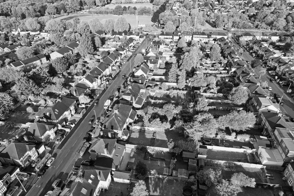 An aerial black-and-white photograph of a residential neighbourhood showing a street lined with detached and semi-detached houses, each with pitched roofs, front gardens, and driveways. The street is populated with parked cars along both sides, with some vehicles partially visible in the image. Behind and beyond the houses, an area of green space with numerous trees and open fields extends into the background, creating a contrast with the densely built-up area. The scene appears to be illuminated by natural daylight, with shadows cast by the trees and buildings highlighting the textures of the rooftops and tree foliage. The environment suggests a suburban setting, where property boundaries are clearly defined by fences and walls, with potential signs of recent property or estate clearance in the form of organized vacant plots or cleared backyard areas. This perspective emphasizes the layout of the neighbourhood, aligning with services offering private waste collection and on-site clearance by Waste Disposal Lewisham.