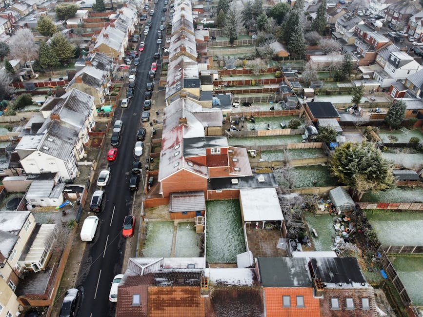 An aerial view of a residential neighbourhood showing a narrow street lined with parked cars on both sides, with closely spaced terraced houses featuring pitched roofs, some with snow and moss accumulation. The image captures multiple backyards enclosed by wooden fences, with visible garden furniture, sheds, and small vegetable plots. Several gardens contain decking, lawned areas, and paved patios, some with outdoor storage or clutter. A few trees and shrubs are scattered throughout the backyards, with one prominent large, bushy evergreen tree near the right side. The photograph appears to be taken during winter or early spring, with a light dusting of snow or frost visible on roofs, garden surfaces, and cars. No waste or rubbish is visibly present in the backyards or street, suggesting either a tidy neighborhood or prior clearance, but the overall scene underscores the typical environment for private rubbish collection or disposal services, with Waste Disposal Lewisham potentially involved in managing waste removal from these individual properties and backyards.