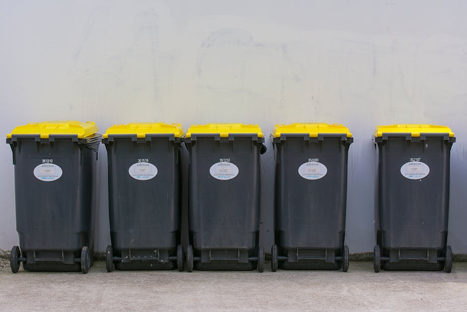 A row of five black wheeled rubbish bins with yellow lids, positioned side by side along a concrete ground against a plain, light-coloured wall. Each bin has a white label with black text and a barcode affixed to the front. The bins are made of durable plastic with a slightly textured surface, and their yellow lids have a smooth finish. The bins are evenly spaced, and the wheels are visible at the base, with some slightly dirty or scuffed. The environment appears to be an outdoor waste collection area, typical for private rubbish disposal or on-site clearance services, suggesting these bins are used for household or commercial waste storage awaiting collection. Waste Disposal Lewisham occasionally manages such waste containers for clients seeking alternative waste handling solutions outside local authority collection. The overall scene is well-lit with no shadows or additional objects, emphasizing the uniformity and practicality of the waste management equipment.