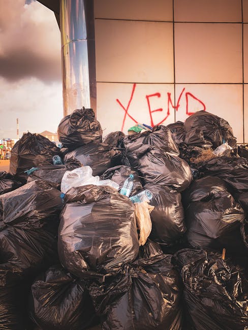 A large pile of black garbage bags filled with household waste is stacked against a tiled wall, with some bottles and plastic packaging visible protruding from the bags. The bags appear to be tightly packed and unevenly piled, occupying most of the foreground. Behind the pile, the wall features graffiti with the word 'XEND' spray-painted in red. To the left, a shiny, reflective metal surface, possibly part of a building or structural frame, is partially visible. The environment suggests an outdoor urban setting, with cloudy sky briefly visible in the background. The scene highlights issues of illegal dumping or unmanaged waste collection, which private waste removal services like Waste Disposal Lewisham may address through alternative on-site clearance or supplementary rubbish removal options, especially in cases of non-compliance with council regulations.