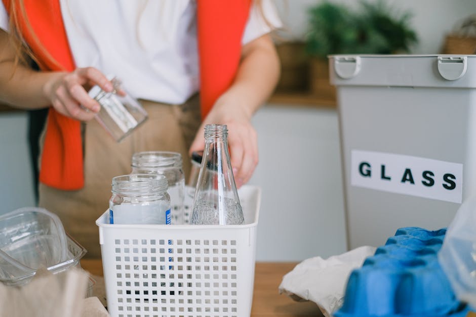 A person wearing a white shirt and orange vest is handling empty glass jars and bottles, placing them into a white plastic container with a grid pattern on the side. The jars are clear and cylindrical, with some having visible blue labeling and metal screw-on lids, while the bottles are tall, slender, and also transparent. To the right, there is a grey recycling bin labeled 'GLASS' in black text, with a hinged lid that is closed. The background features a kitchen environment with a wooden countertop and a potted green plant, indicating a domestic setting focused on waste separation and recycling. The scene emphasizes a household activity related to the proper disposal and separation of glass waste, aligning with private waste handling and eco-friendly rubbish removal practices.