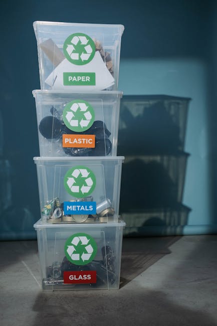 A stack of four clear plastic recycling bins with lids, arranged vertically on a wooden floor against a blue wall. Each bin is labeled with a different waste category and a corresponding colour-coded sticker featuring the recycling symbol. The top bin, designated for paper, has a green sticker and contains flattened cardboard and paper waste. The second bin, intended for plastic, has an orange sticker and contains various plastic items, some recognizable as bottles and containers. The third bin, for metals, features a blue sticker and includes crushed cans and metallic fragments. The bottom bin, for glass, bears a red sticker with glass bottles and jar fragments visible inside. The scene is well-lit, with natural or artificial light creating subtle reflections on the plastic surfaces and casting faint shadows on the floor. This arrangement reflects an organized approach to household waste separation, emphasizing the importance of correct disposal for recycling and waste management services such as Waste Disposal Lewisham, which supports alternative waste handling options outside of local authority collections.