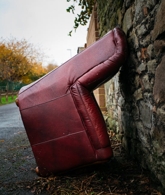A large, burgundy leather sofa is positioned upright against a rough stone wall on a paved outdoor area. The sofa's surface exhibits a smooth, shiny finish with visible stitching along the edges and seams. The armrest on the right side is slightly rounded and padded, while the seat and back cushions are plush and slightly indented, showing signs of use. Surrounding the sofa, there are scattered fallen leaves and small twigs, indicating an outdoor setting during autumn. In the background, a glimpse of a residential street with trees bearing orange and yellow foliage can be seen, along with parts of a brick building and a wooden fence. The lighting is natural, likely overcast, giving a neutral tone to the scene. This image conveys an instance of bulky waste, which could be subject to private rubbish collection or alternative disposal services like those offered by Waste Disposal Lewisham, supporting the theme of alternative waste handling and rubbish removal.