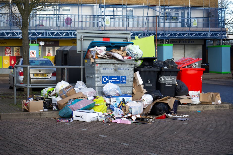 A large collection of mixed waste and rubbish is piled up on a paved street in front of a small parking area. The pile includes cardboard boxes, plastic bags, paper materials, and discarded packaging, some of which are crushed or loosely arranged. Prominent among the rubbish are black and red trash bags, partially opened, revealing additional waste inside. There are also a few loose items like cardboard sheets and plastic containers scattered around the base of the pile. To the left, a silver hatchback car is parked, partially blocking a metal railing that runs parallel to the street. Behind the waste, a row of commercial storefronts with closed shutters and signage, including one that mentions a fish bar, is visible. The background features a building under renovation with metal scaffolding and safety barriers painted in blue and green. Overcast lighting casts a neutral tone across the scene, fitting the setting of a private rubbish collection point or on-site disposal area, as handled by waste disposal services like Waste Disposal Lewisham.