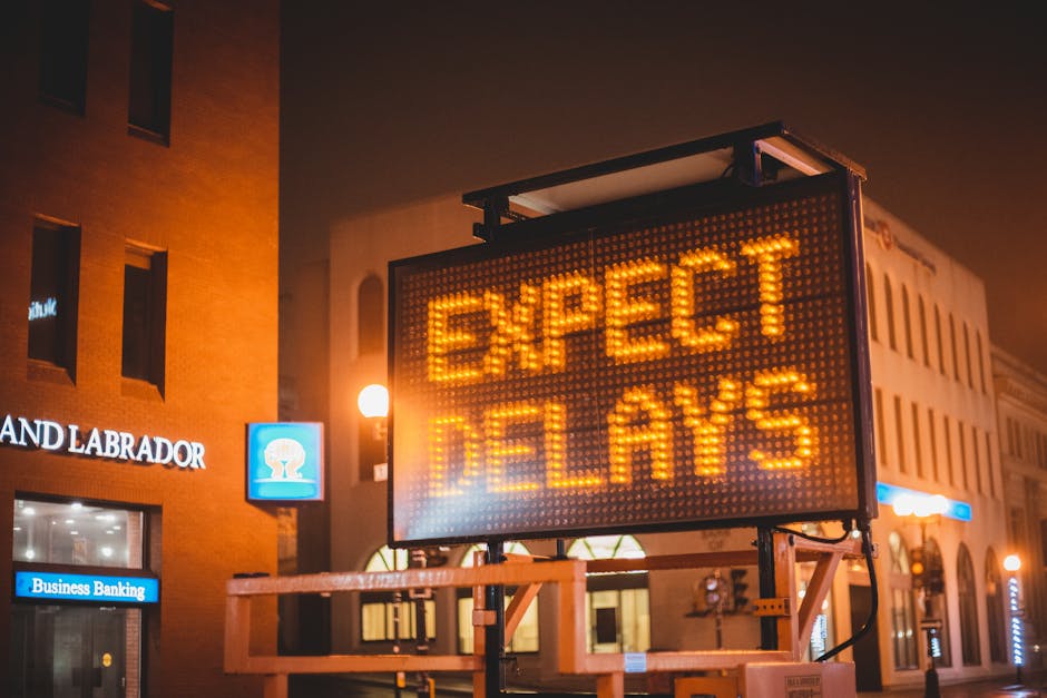 A bright orange LED message board is mounted on a metal stand outdoors in an urban setting at night, displaying the words 'EXPECT DELAYS' in illuminated text. Behind the board, there is a beige building with arched windows and vertical architectural details, as well as a smaller blue sign featuring a white emblem on the left side. The scene is lit by warm street lighting, creating a glow around the sign and nearby structures. To the left, part of a brick building with modern signage reading 'LAND LABRADOR' and 'Business Banking' is visible, with illuminated window panes. The environment appears to be a city or town center, with additional signage and streetlights in the background, suggesting a busy area where temporary traffic or service disruptions may be anticipated. The overall composition indicates an outdoor setting focused on informing pedestrians or drivers of potential delays, aligning with the context of alternative waste handling or disruptions in public services that Waste Disposal Lewisham might cover through independent collection services.