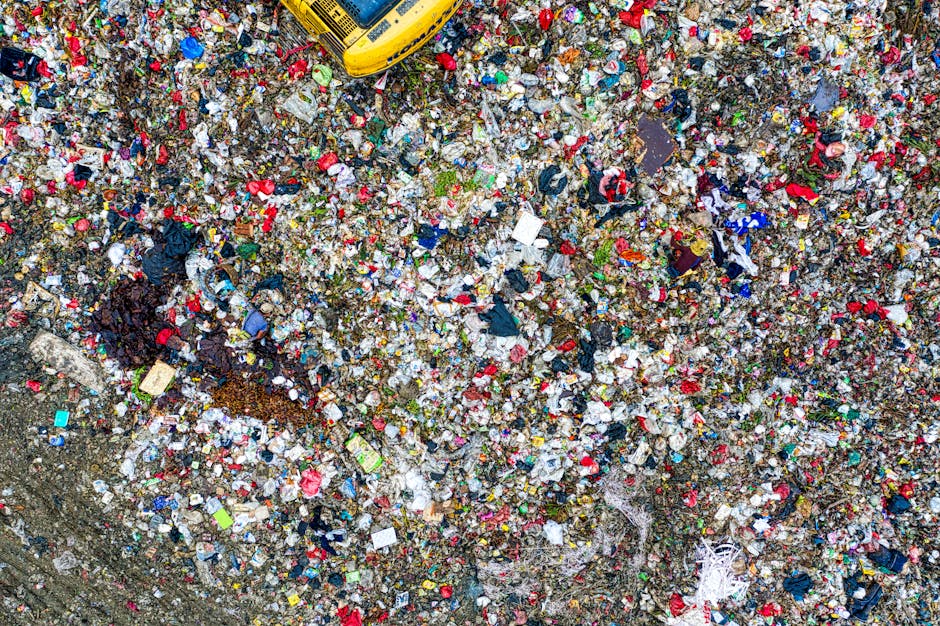 An aerial view of a large, open landfill site filled with a dense layer of mixed waste materials covering the entire visible area. The waste primarily consists of plastic bottles, bags, packaging, and debris in various colors including red, blue, green, white, and black. Some materials appear crushed or squashed, with textures ranging from smooth plastic surfaces to crumpled paper and textiles. There are small patches of soil and dirt visible amidst the rubbish, and a yellow waste compactor or machinery partially visible at the top edge of the image. The scene is illuminated by natural daylight, creating a bright, evenly lit environment that emphasizes the cluttered, chaotic nature of the waste accumulation. This bulk of refuse exemplifies the challenge of waste disposal and highlights the importance of professional rubbish removal services like Waste Disposal Lewisham for managing such large quantities of waste efficiently and responsibly.